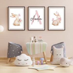 May include: Three framed prints in a pink and white colour scheme. The left print features a watercolour illustration of a brown bunny with a pink flower crown and a butterfly. The middle print features a pink letter A with the name Amelia written in black script. The right print features a watercolour illustration of a brown bunny with a pink flower crown and a pink bow.