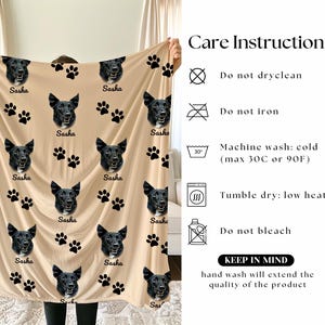 a person holding up a beige blanket with black paw prints and the word "Sasha" printed on it. The blanket has a care instruction label attached to it, providing guidelines for washing and care.