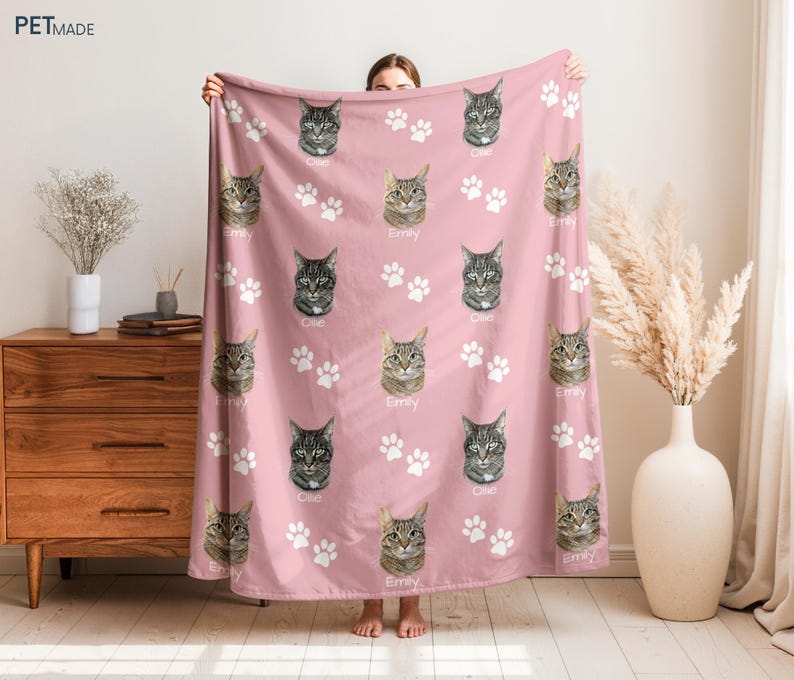 A person is holding up a pink blanket with cat faces and paw prints on it, which is placed on a wooden dresser.