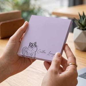 May include: A lilac-coloured notepad featuring a black and white cat illustration and the name "Caitlin Leary" in cursive script. The notepad is held in someone's hands.