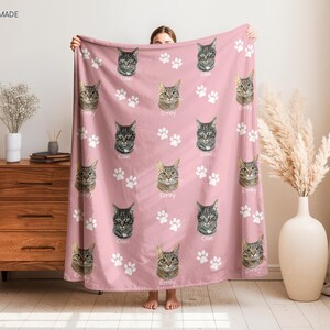 A person is holding up a pink blanket with cat faces and paw prints on it, which is placed on a wooden dresser.