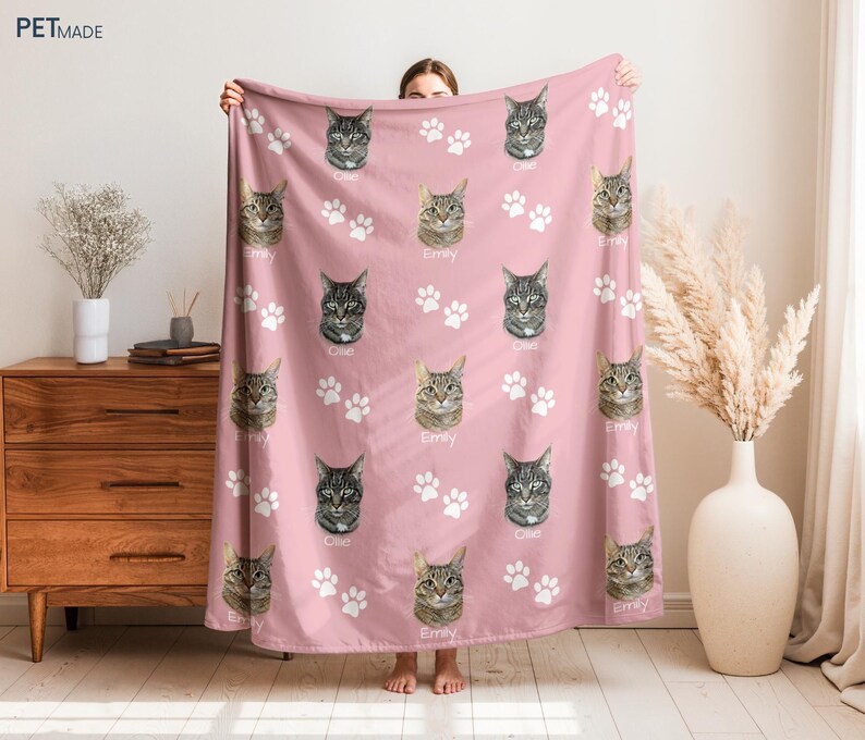 A person is holding up a pink blanket with cat faces and paw prints on it, which is placed on a wooden dresser.