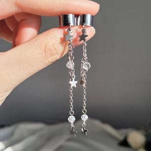 May include: Silver-toned ear cuffs with dangling chains. Each chain features small star charms and clear, round beads. The jewelry is held up by a hand against a soft, blurred background.