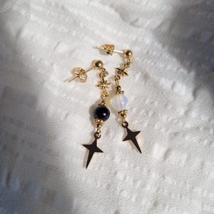 May include: A pair of gold-tone drop earrings featuring a stud post, a small star charm, and a dangling bead. One earring has a black bead, and the other has a translucent white bead, both finished with a four-pointed star charm at the bottom.