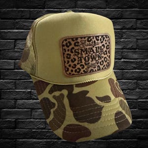 May include: A khaki trucker hat with a camouflage brim. The hat has a brown leather patch with the text "Just a Small Town girl" in a leopard print design.