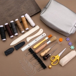 May include: A leather craft kit with tools and supplies, including needles, thread, a thimble, a ruler, a brush, a small bottle, a pair of scissors, and a silver zippered pouch.