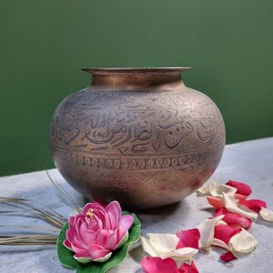 May include: A large, round, brass pot with intricate Arabic calligraphy etched into the surface. The pot is sitting on a white surface with pink and white flower petals scattered around it.