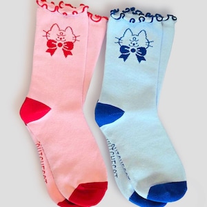 Könnte beinhalten: Ein Paar Crew-Socken, eine rosa und eine hellblaue, mit einem Katzen-Gesicht-Design. Die rosa Socke hat ein rotes Katzen-Gesicht und eine Schleife, und die blaue Socke hat ein blaues Katzen-Gesicht und eine Schleife. Beide Socken haben einen gerüschten oberen Rand.