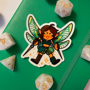 May include: A sticker featuring a cartoon fairy with brown hair, green wings, and a green outfit, holding two daggers. The sticker is on a green surface, surrounded by white and gold dice.