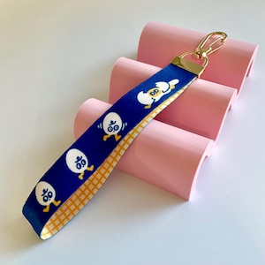 Puede incluir: Una correa de muñeca azul con un cierre dorado y un patrón a cuadros amarillo y blanco en la parte inferior. La correa presenta un patrón repetido de huevos blancos con piernas y brazos amarillos sobre un fondo azul.