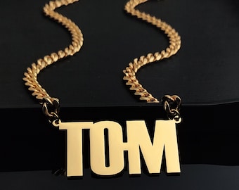 Puede incluir: Collar de cadena dorada con un colgante de oro que dice "TOM" en letras mayúsculas.