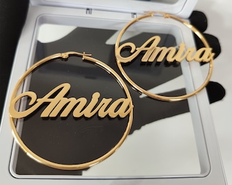 May include: A pair of gold hoop earrings with the name "Amira" written in cursive on each earring.