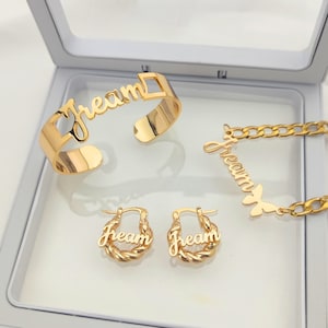 Puede incluir: Conjunto de joyas doradas con la palabra "Dream" en cada pieza. El conjunto incluye un brazalete, un collar y un par de pendientes.