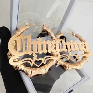 May include: Gold hoop earrings with the word "Thingthing" in a gothic font. The earrings are made of bamboo-style metal.