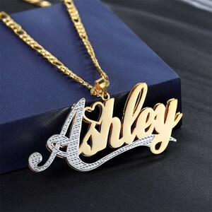 Puede incluir: Collar de tono dorado y plateado con un colgante de nombre en cursiva que dice "Ashley". El colgante tiene un pequeño corazón en la "A".