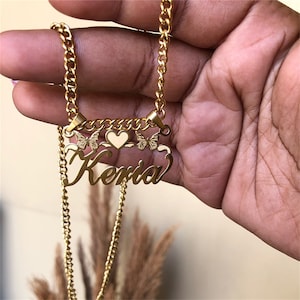 K&ouml;nnte beinhalten: Goldkette mit einem Anh&auml;nger mit dem Namen "Keria" in einer kursiv geschriebenen Schrift, verziert mit zwei Schmetterlingsanh&auml;ngern und einem Herzanh&auml;nger.