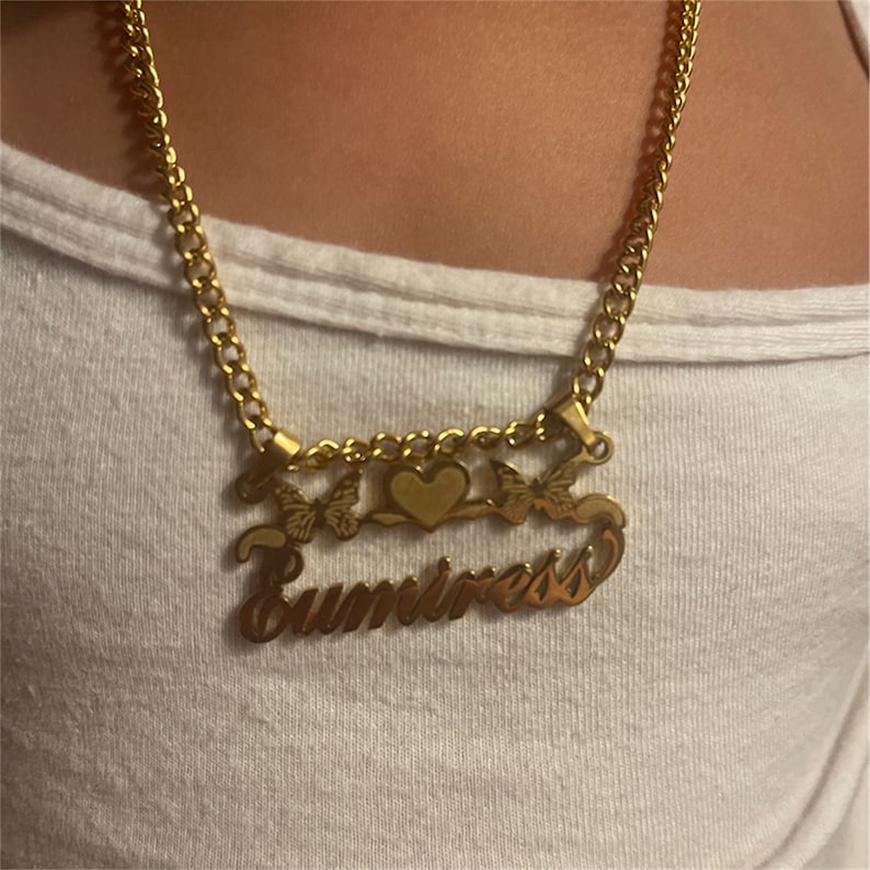 K&ouml;nnte beinhalten: Goldkette mit einem Herz-Anh&auml;nger und zwei Schmetterlings-Anh&auml;ngern. Die Kette hat einen Namensschild-Anh&auml;nger mit der Aufschrift "Sumiress".