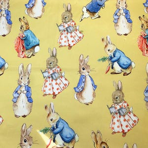 May include: A yellow fabric featuring a repeating pattern of watercolor-style rabbits. Some rabbits wear blue jackets, while others wear white dresses with red polka dots. Several rabbits are depicted holding books or carrots in various poses.