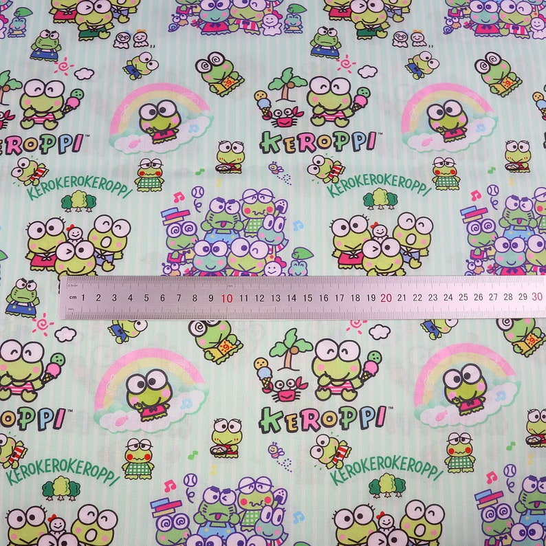 May include: Green fabric with a repeating pattern of a cartoon frog character named Keroppi. The fabric has a light green background with white stripes and features a rainbow, palm trees, and other cartoon elements. The text "Keroppi" and "Kerokerokeroppi" appears on the fabric.