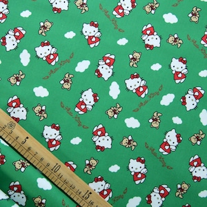 Puede incluir: Tela verde con un patrón repetido de personajes de Hello Kitty con vestidos y lazos rojos, pequeños osos de peluche marrones y nubes blancas. Las palabras "Hello Kitty" están escritas en cursiva. Una regla de madera está en la imagen.