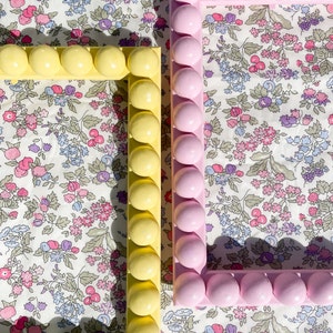 May include: Two decorative picture frames with a floral pattern background. The frame on the left is yellow with round embellishments. The frame on the right is pink with round embellishments.