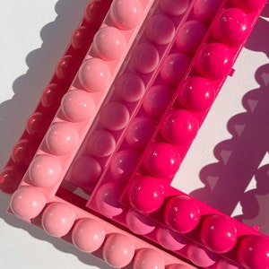 May include: A pink, red, and magenta frame made of a textured plastic with a series of raised circles. The frame is made of three layers of plastic, each a different shade of pink.