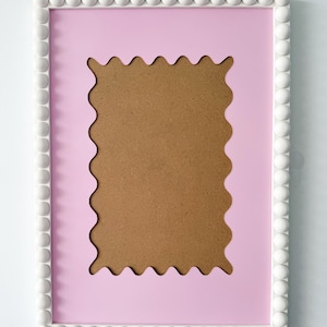 Puede incluir: Marco de fotos rectangular con interior rosa claro y borde de cuentas blanco. El marco tiene un borde festoneado alrededor del interior rosa y la parte posterior es de cartón marrón.