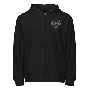 May include: Black zip-up hoodie with a hood and a front pocket. The hoodie features a circular emblem on the upper left chest with a stylized bird design in shades of gray and white, set against a dark purple background.