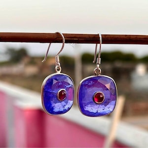 May include: A pair of silver earrings featuring square, blue gemstones with a central red stone. The earrings have silver hooks. The gemstones are faceted.