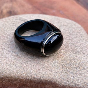 May include: A polished black ring featuring a rounded, oval-shaped stone set within a silver band. The ring, made from a dark, glossy material, rests on a light brown stone surface. The design of the ring is simple and elegant.