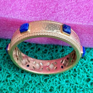 K&ouml;nnte beinhalten: Ein goldfarbenes Armband mit strukturierter Oberfl&auml;che und aufwendigen Details. Das Armband hat quadratische, tiefblaue Edelsteine, die in regelm&auml;&szlig;igen Abst&auml;nden angeordnet sind. Der Hintergrund ist eine Mischung aus Rosa und Gr&uuml;n.
