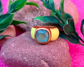 May include: A bold ring featuring a gold band and a square, pale blue gemstone. The ring's band is a vibrant yellow. The ring is presented on a grey stone, with green leaves and a pink backdrop.