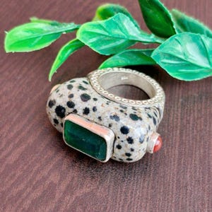 May include: A unique ring featuring a speckled stone base with black spots, a silver band, and a rectangular green gemstone. A small red stone accents the side. The ring is a statement piece.