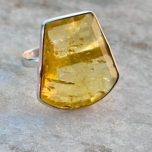 May include: A silver ring with a large, faceted, yellow gemstone. The gemstone has an unusual, irregular shape and is set in a simple silver band. The ring is shown against a neutral, textured background, emphasising the stone's colour and clarity.