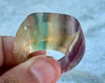 Puede incluir: Un anillo de fluorita multicolor con forma rectangular redondeada. El anillo está hecho de una piedra translúcida con tonos de verde, amarillo, morado y blanco.