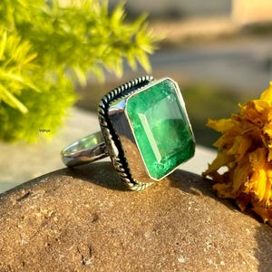 Puede incluir: Un anillo de plata con una gran gema verde rectangular con talla esmeralda. El anillo tiene un engaste detallado y texturizado alrededor de la esmeralda. El anillo se exhibe sobre una piedra marrón, con flores amarillas y follaje verde en el fondo.