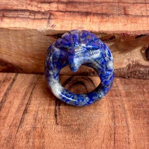 Puede incluir: Un anillo tallado en lapislázuli con un diseño de cabeza de águila estilizada. El anillo es de color azul oscuro con vetas blancas y grises. El anillo está sobre un fondo de madera.