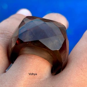 May include: A large, faceted, dark brown ring with a geometric design. The ring is worn on a finger, with the word "Vidhya" visible. The ring's color is a deep, translucent brown, and it has a bold, angular shape.