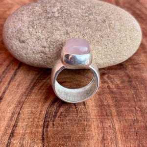 Könnte beinhalten: Ein silberner Ring mit einem großen, hellrosa, quadratisch geschliffenen Edelstein. Der Ring liegt auf einer Holzoberfläche mit einem großen, glatten, grauen Stein im Hintergrund. Der Ring hat ein dickes Band mit gebürsteter Metalloberfläche.