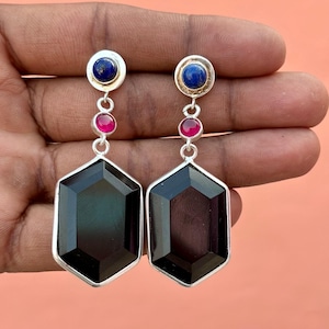 Könnte beinhalten: Ein Paar silberne Ohrringe mit geometrischem Design. Jeder Ohrring hat einen dunklen, facettierten, sechseckigen Stein, einen kleinen, runden, magenta-farbenen Stein und einen blauen Stein oben. Die Ohrringe werden in einer Hand vor einem pfirsichfarbenen Hintergrund gehalten.