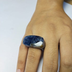 以下が含まれることがあります： ファセットデザインの大きな濃い青色のサファイアリング。リングはシルバーのバンドにセットされており、指につけられています。