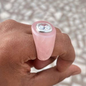 May include: A pink rose quartz ring with a square, clear gemstone set in a silver bezel. The ring is a light pink color and has a smooth, polished finish. The gemstone is clear and sparkling.