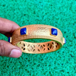 K&ouml;nnte beinhalten: Goldfarbenes Armreif-Armband mit geh&auml;mmertem Muster und quadratischen, tiefblauen Edelsteinen. Das Armband hat dekorative Ausschnitte und einen gemusterten Rand. Ein Schmuckst&uuml;ck.