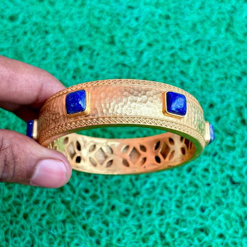 K&ouml;nnte beinhalten: Goldfarbenes Armreif-Armband mit geh&auml;mmertem Muster und quadratischen, tiefblauen Edelsteinen. Das Armband hat dekorative Ausschnitte und einen gemusterten Rand. Ein Schmuckst&uuml;ck.