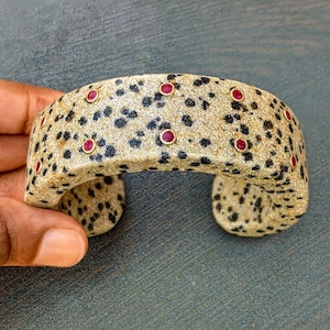 May include: A beige cuff bracelet with black spots and red gemstone accents. The bracelet has a speckled pattern and is adorned with small, round red gemstones set in gold-coloured bezels. A unique piece of jewellery.