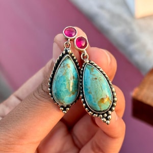 May include: A pair of silver dangle earrings with turquoise stones and pink accents. The turquoise stones are teardrop shaped and have a marbled pattern. The pink accents are small round stones.