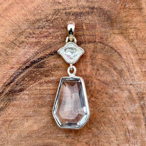 Pu&ograve; includere: Un ciondolo con pietra preziosa trasparente e sfaccettata con montatura in argento. Il ciondolo presenta una grande pietra a forma di goccia sotto una pietra pi&ugrave; piccola a forma di diamante. Il gioiello &egrave; esposto su una superficie di legno.