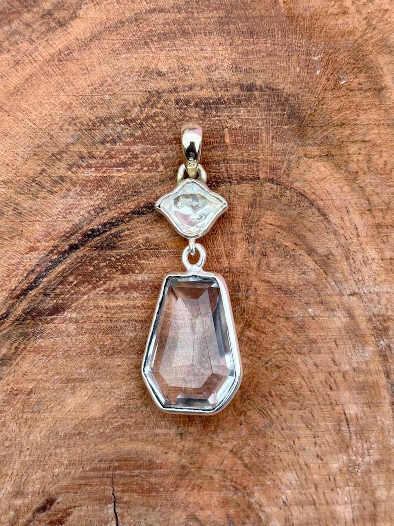 Pu&ograve; includere: Un ciondolo con pietra preziosa trasparente e sfaccettata con montatura in argento. Il ciondolo presenta una grande pietra a forma di goccia sotto una pietra pi&ugrave; piccola a forma di diamante. Il gioiello &egrave; esposto su una superficie di legno.