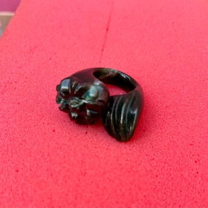 Puede incluir: Anillo verde oscuro con un diseño floral detallado en un lado y una forma curva texturizada en el otro. El anillo se encuentra sobre un fondo rojo brillante, resaltando sus intrincados detalles y su forma única.