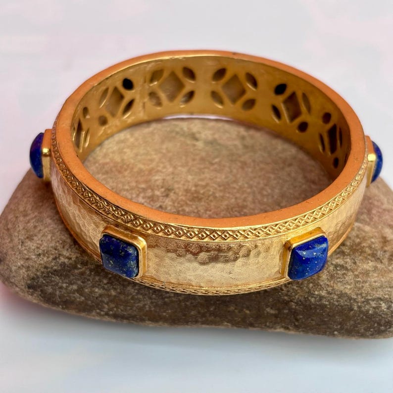 K&ouml;nnte beinhalten: Ein goldfarbenes Armreif-Armband mit geh&auml;mmerten Texturen und geometrischen Aussparungen. Das Armband hat drei quadratische, tiefblaue Edelsteine. Das Armband liegt auf einer Steinoberfl&auml;che.